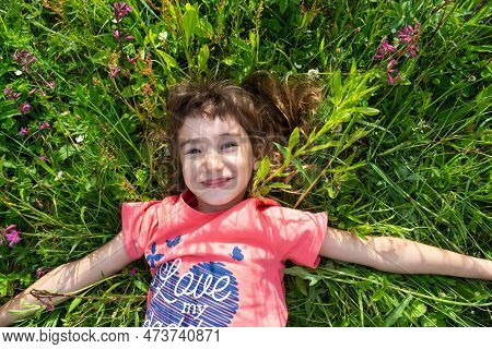 Portrait Of A Child Girl In Summer Lying In The Grass And Wildflowers With Heels And Palms. Summer T