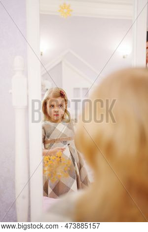 A Cute Little Girl In A Plaid Dress Looks In The Mirror And Primping.