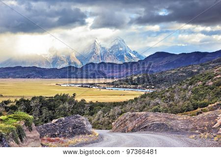 The road between turned yellow hills and river goes to snow-covered black rocks. Patagonia. Vertiginous landscape in the Chilean Andes