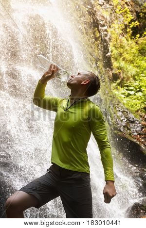 hiker man drinking fresh clean water from a natural. Young tourist drinking water from a bottle outdoor waterfall in the background. Drink water, hydration, healthy lifestyle, hiking concept