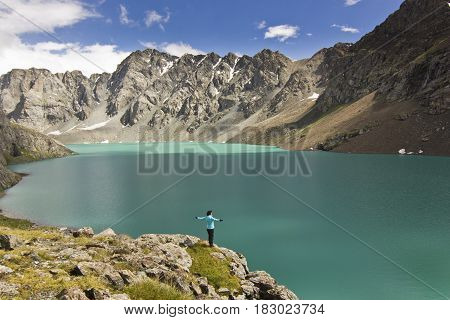 girl in blue jacket standing with hands up above blue calm mountain lake Alakol with mountains surrounded