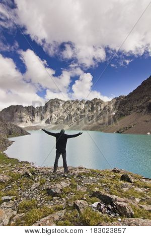 man in black jacket standing with hands up above blue calm mountain lake Alakol with mountains surrounded