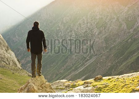 man in black jacket standing with hands up above mountain slopes at yellow sunset