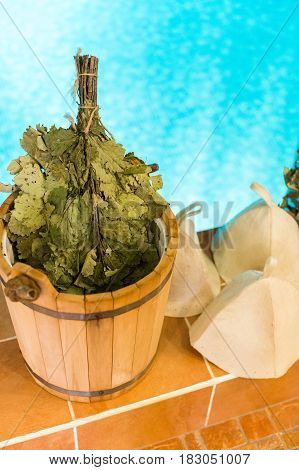 Oak Leaf Venik With Dried Herbs in wooden sauna bucket and sauna hat on a pool background. Medical sauna broom and health Venik for body. Russian banya and sauna accessories. relaxation concept.