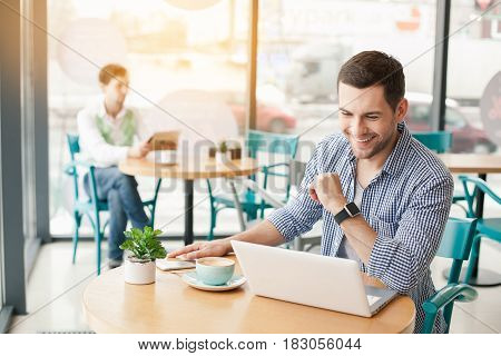 Coffee time. Handsome young man in cafe with big window. Man with cup of coffee. Man using laptop and smiling