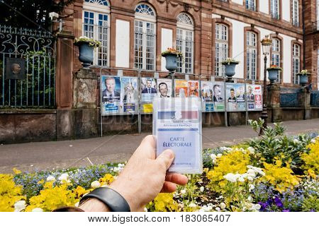 STRASBOURG FRANCE - APR 23 2017: Man holding Carte Electorale - voter's card French voter registration card in front of official campaign posters for all eleven candidates for the 2017 French presidential elections outside pooling station