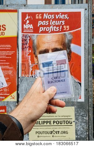 STRASBOURG FRANCE - APR 23 2017: French voter registration card held by male hand in front of official campaign poster of Philippe Poutou candidate for the 2017 French presidential elections posted outside a polling station