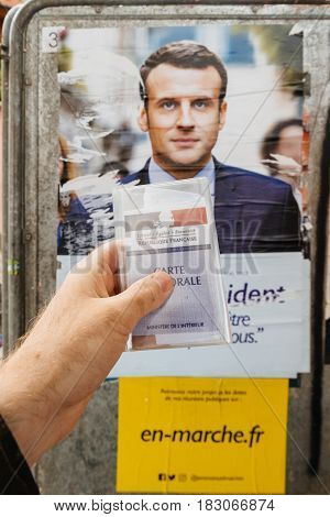 STRASBOURG FRANCE - APR 23 2017: French voter registration card held by male hand in front of official campaign poster of Emmanuel Macron candidate for the 2017 French presidential elections posted outside a polling station