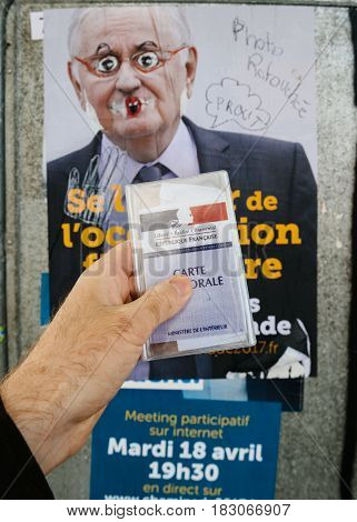 STRASBOURG FRANCE - APR 23 2017: French voter registration card held by male hand in front of official campaign poster of Jacques Cheminade candidate for the 2017 French presidential elections posted outside a polling station