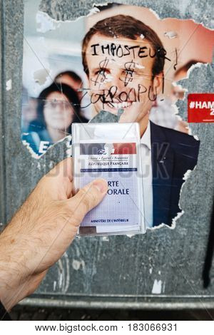 STRASBOURG FRANCE - APR 23 2017: French voter registration card held by male hand in front of official campaign poster of Emmanuel Macron candidate for the 2017 French presidential elections posted outside a polling station