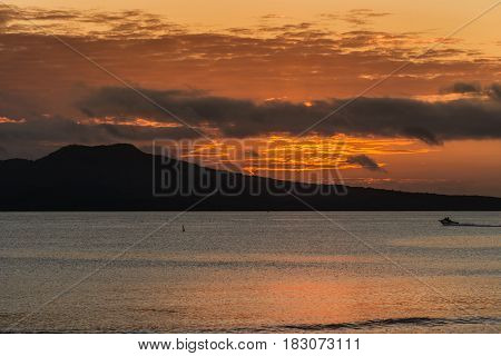 Auckland New Zealand - March 2 2017: Sunrise over Rangitoto Volcano shot from Tapapuna Beach in the Hauraki Gulf. Shot 1 of 4. Fire lines in sky among dark clouds. Black volcano. Blue-gray ocean water reflects orange sky.