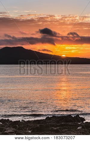 Auckland New Zealand - March 2 2017: Sunrise over Rangitoto Volcano shot from Tapapuna Beach in the Hauraki Gulf. Shot 2 of 4. Half sun Black volcano. Blue-gray ocean water with orange reflections