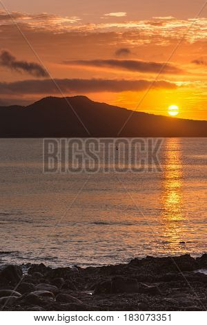 Auckland New Zealand - March 2 2017: Sunrise over Rangitoto Volcano shot from Tapapuna Beach in the Hauraki Gulf. Shot 4 of 4. Full sun Black volcano. Blue-gray ocean water with orange reflections.
