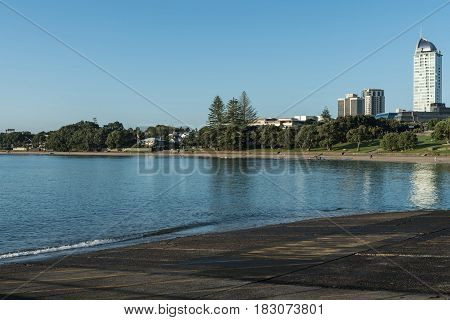 Auckland New Zealand - March 2 2017: Black volcanic south shore of Tapapuna Beach shows park highrise and canoe rowers under blue sky with reflecting sea water.