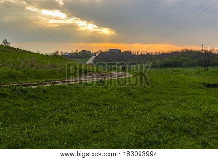 Field and dirt road on the hilly terrain leading to the village.Above the village a gloomy sky with the gleams of the morning Sun.On the road is the figure of a man.