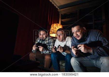 Three high concentrated young guys with joysticks play exciting video game at home in dark room. Tension to win tourney.
