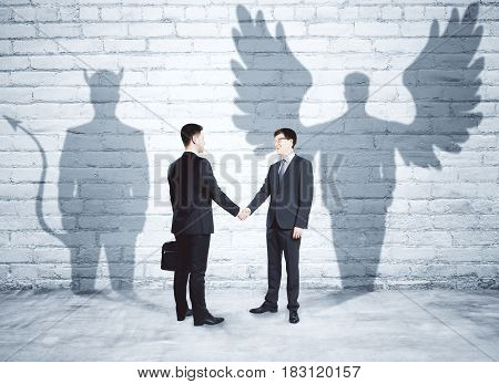 Businessmen with angel and demon shadows shaking hands in brick interior. Team work concept