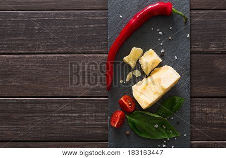 Cheese delikatessen closeup on black stone desk at wooden surface background. Parmesan pieces decorated with chili pepper and cherry tomatoes, top view image with copy space