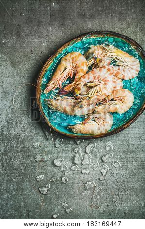 Raw uncooked tiger prawns on chipped ice in turquoise blue tray over grey concrete background, top view, copy space, vertical composition. Fresh seafood