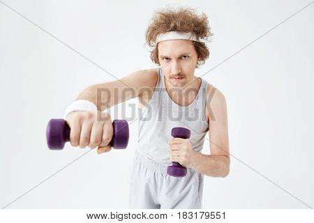 Young concentrated man working on arm muscles, training hard with dumbbells. Retro style. Looking straight