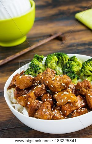 Chicken lacquered with a sweet soy teriyaki sauce in a white bowl. Garnished with rice and broccoli. Chopsticks brown wooden table.