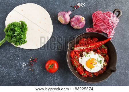 Huevos Rancheros traditional mexican dish fried eggs and salsa on a black cast-iron skillet on dark concrete background top view.