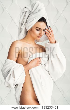 sexy young woman with dark hair, big eyes and dark eyebrows wearing white bath robe whith towel on her head. Model with light nude make-up, white studio background, beauty photo