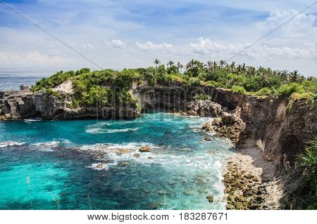 iew of clear blue lagoon and cliff side with palm trees in the distance. Ceningan Island, Bali.