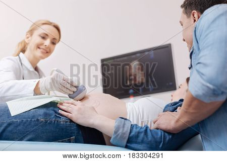 Admiring first digital image of our child. Smiling mature skillful medic working in the sonography cabinet while monitoring pregnant belly and using ultrasound linear transducer
