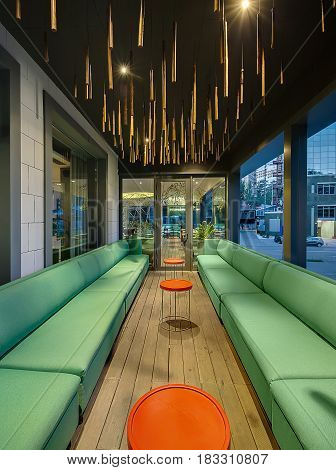 Hall with large windows and luminous lamps in the modern cafe. There are green sofas and round orange tables between them. On the ceiling there are many hanging wooden decorations. Vertical.