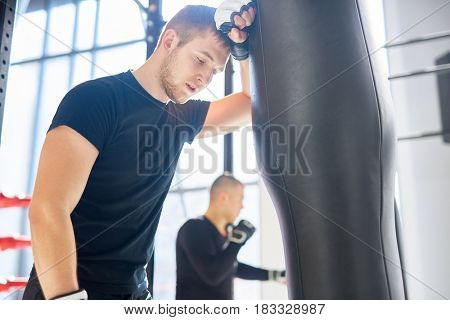 Low angle portrait of young tired sports man leaning on punching bag during boxing practice in fight club