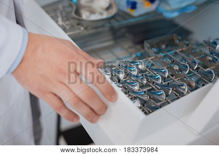Take it. Young doctor wearing white smock putting right hand on the shelf while choosing ear-funnel