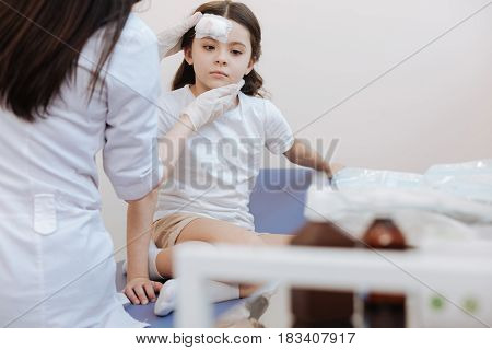 Look at me. Pleasant nice female doctor holding her patients head and checking the forehead wound while sitting in front of her