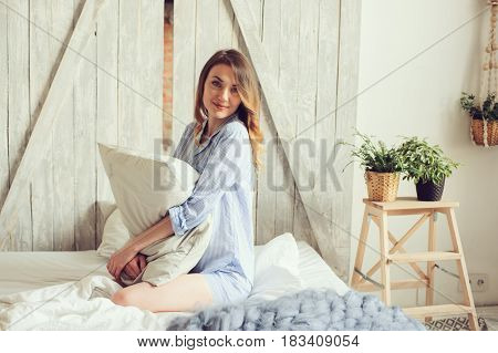 young woman in pajama wake up in the morning in cozy scandinavian bedroom and sitting on bed with white bedlinen. Casual lifestyle in modern interior