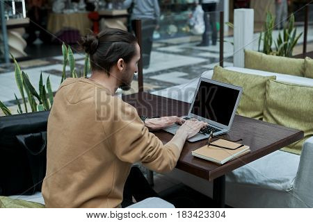 Young journalist working in open space in shopping center. Man typing message for a friends in the internet social network on mock up notebook. Back view on freelancer develop on new IT application