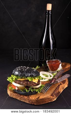 Fresh homemade burger on wooden serving board with fork and knife tomato sauce and vitage bottle on dark background.