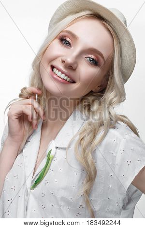 Young smiling girl with curls in a hat. A beautiful model in a white shirt and a brooch-flower. Gentle makeup. Summer light image. Beauty of the face.