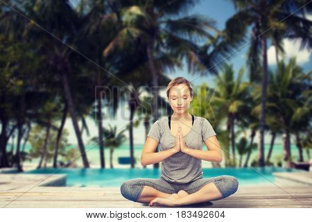 fitness, sport, people, resort and healthy lifestyle concept - woman making yoga meditation in lotus pose over swimming pool and tropical beach with palm trees background