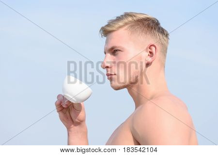 young handsome man with sexy muscular athletic strong body has bare torso and strong belly holding cup or mug of coffee tea with serious face on blue sky background