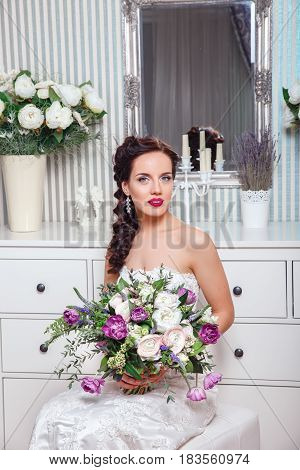 Wedding. Bride in beautiful dress sitting on sofa indoors in white studio interior like at home. Trendy wedding style shot in full length.Young attractive caucasian brunette model like a bride