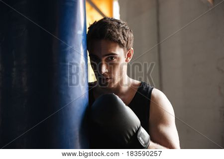 Portrait of strong young boxer near punchbag in shadow in gym