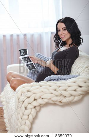 happy beautiful bruntte pregnant woman relaxing on sofa with echo scan in hands