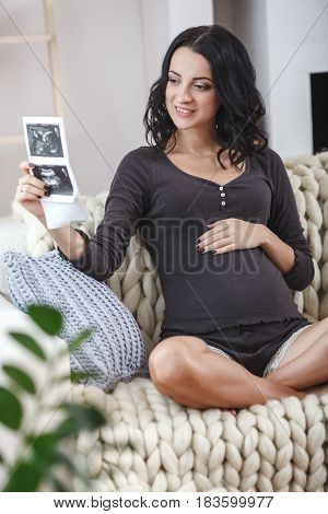 happy beautiful bruntte pregnant woman relaxing on sofa with echo scan in hands