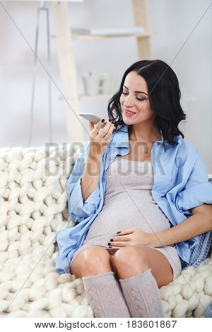 happy pregnant brunette woman using voice command recorder or calling on smartphone at home, she sitting on couch