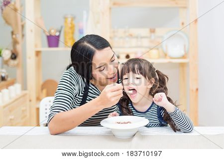 Asian young mother feeding breakfast for her daughter with a kitchen background.