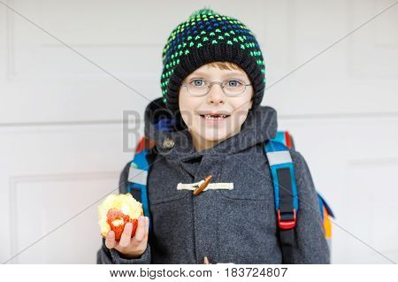 Little kid boy with eye glasses on the way to the school and eating fresh apple. Child with missing front teeth wearing warm clothes, glasses and backpack. Happy student or people. Education concept