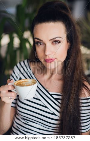 Close up portrait of beautiful but a bit tired woman holding a cup of cappuccino. Blurred background.