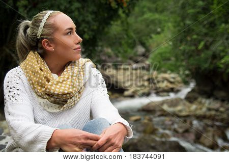 Happy young girl sitting by the river rocks in a forest enjoying the quietness