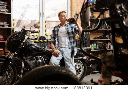 Cheers to craftsmen. Diligent trained virile guy finishing his workday in the repair shop by relaxing on a motorbike he currently repairing