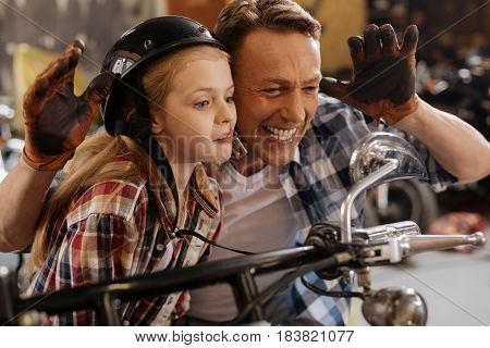 Hilarious couple. Positive artistic cute dad and daughter making funny faces and looking at their reflection while sitting on a bike in garage